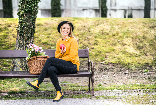 Portrait of a young beautiful woman with flowers in a basket in town. A girl sitting on a bench, eating an apple. Sunny spring.