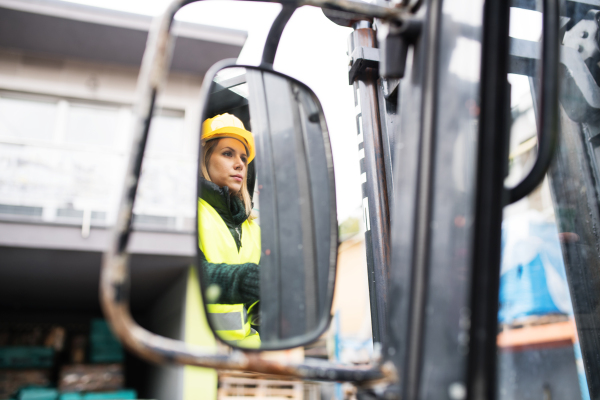 Female forklift truck driver in an industrial area. A woman sitting in the fork lift outside a warehouse. Reflection in a rear view mirror.