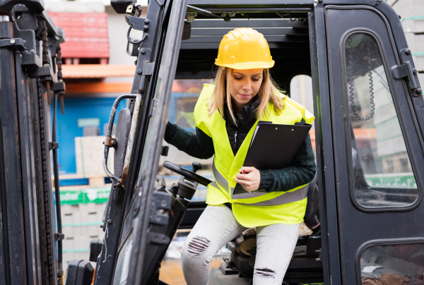 Female forklift truck driver outside a warehouse. A woman getting out of the fork lift truck, holding notes.