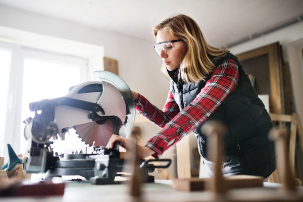 Young woman worker in workroom. Female carpenter using miter saw.