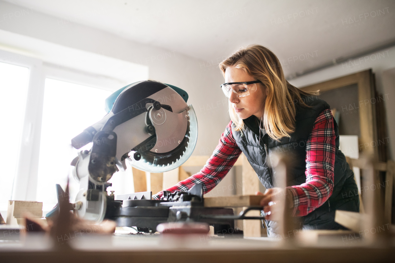 Young woman worker in workroom. Female carpenter using miter saw.