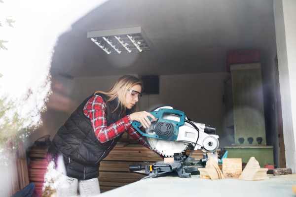 Young woman worker in workroom. Female carpenter using miter saw. Shot through glass.