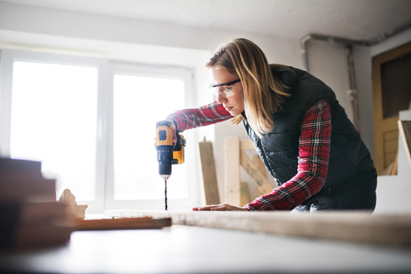 Young woman worker in workroom. Female carpenter using drill.