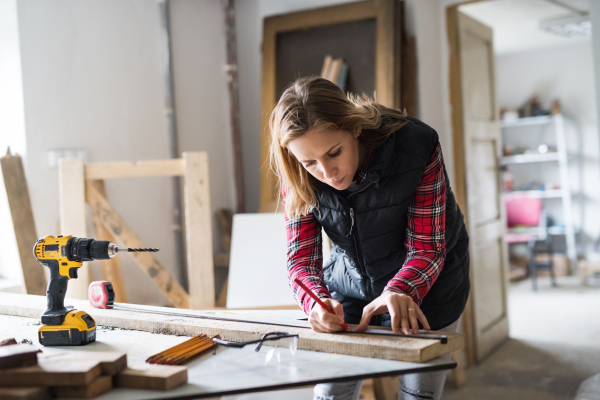 Young woman worker in workroom. Female carpenter measuring wood plank.