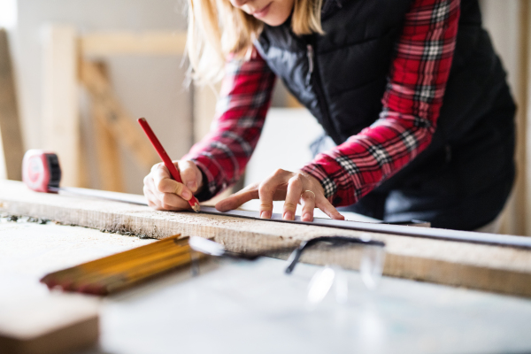 Unrecognizable young woman worker in workroom. Female carpenter measuring wood plank.