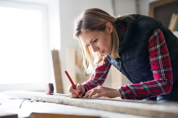 Young woman worker in workroom. Female carpenter measuring wood plank.