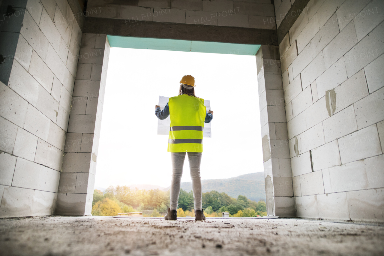 Female worker on the building site. Beautiful young woman holding blueprints. House construction. Rear view.