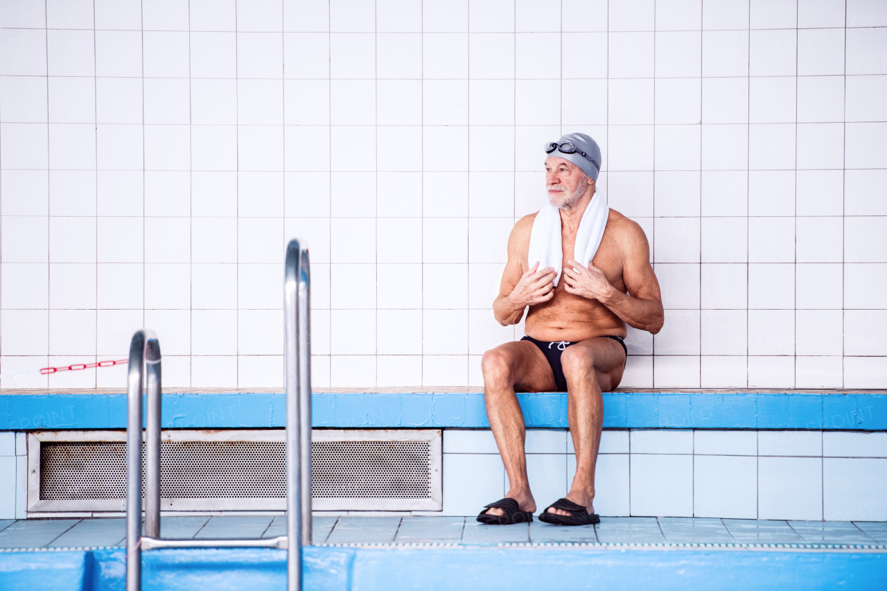 Senior man sitting by the indoor swimming pool, towel around his neck. Active pensioner enjoying sport.