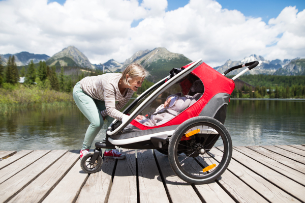 Senior woman and children in jogging stroller at he lake, summer day. High mountains in the background.