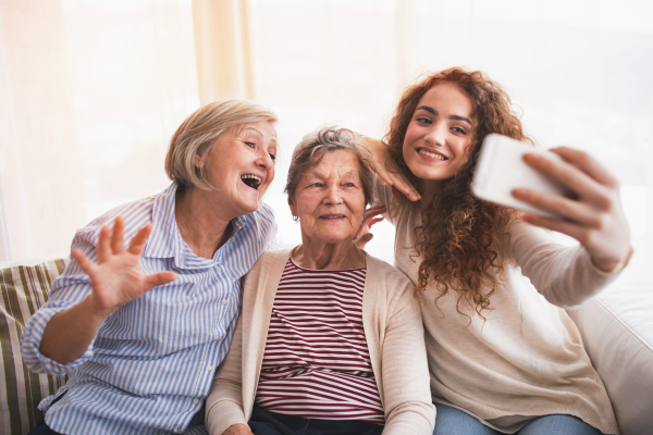 A teenage girl, her mother and grandmother with smartphone at home, taking selfie. Family and generations concept.