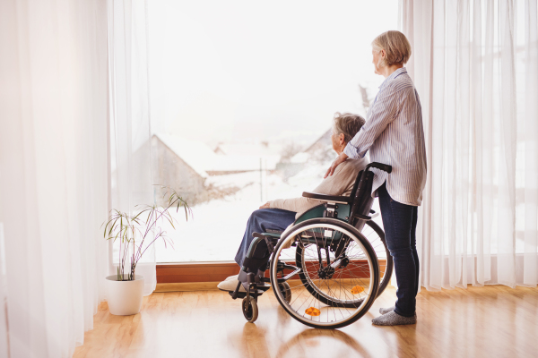 An elderly mother with her daughter at home looking out of the window. A carer assisting a disabled senior woman in wheelchair.