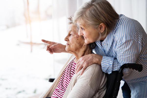 An elderly mother with her daughter at home looking out of the window. A carer assisting a disabled senior woman in wheelchair.