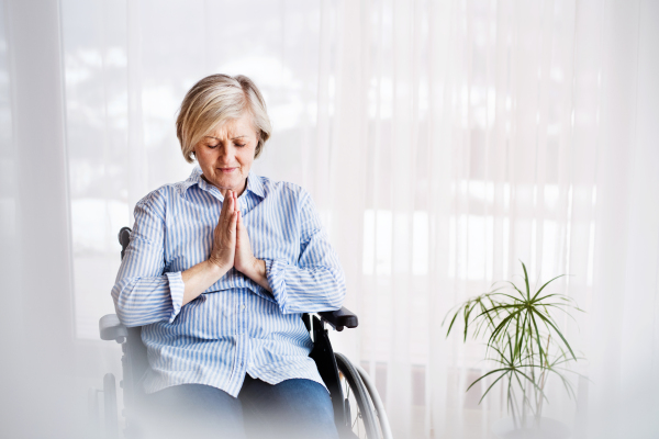 A senior woman in wheelchair praying at home, eyes closed.