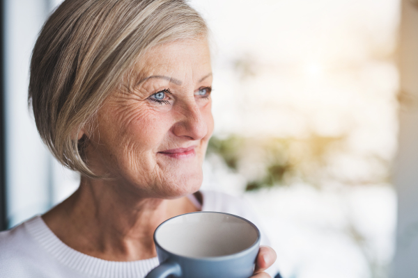 Senior woman in the kitchen. An old woman inside the house, holding a cup of coffee. Close up.