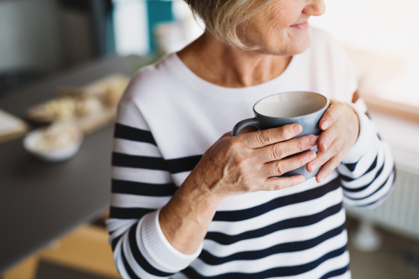 Unrecognizable senior woman in the kitchen. An old woman inside the house, holding a cup of coffee.