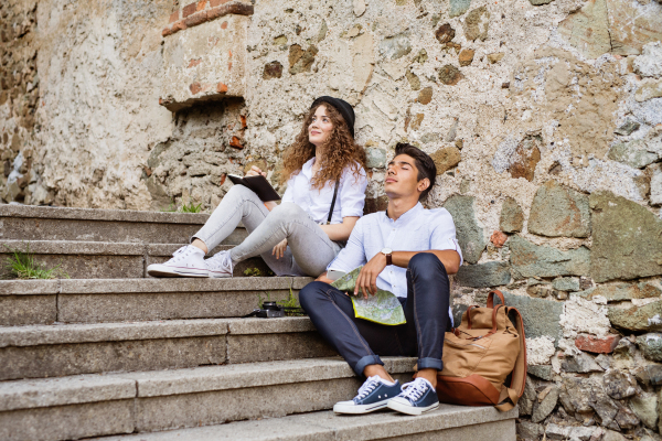 Two beautiful young tourists with map in the old town. Teenagers sitting on stairs, resting.