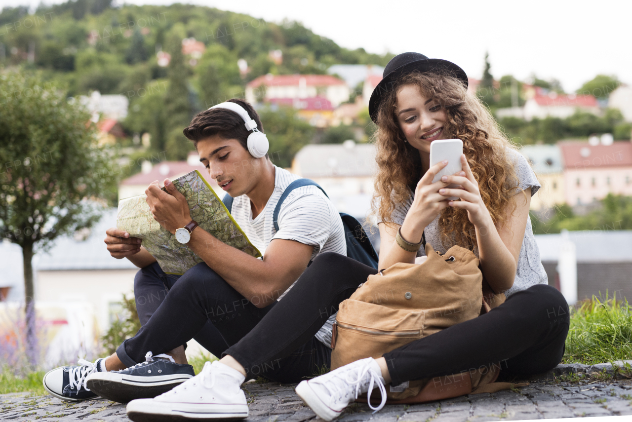 Two beautiful young tourists with smartphone, map and headphones in the old town. Teenagers sitting on the ground.