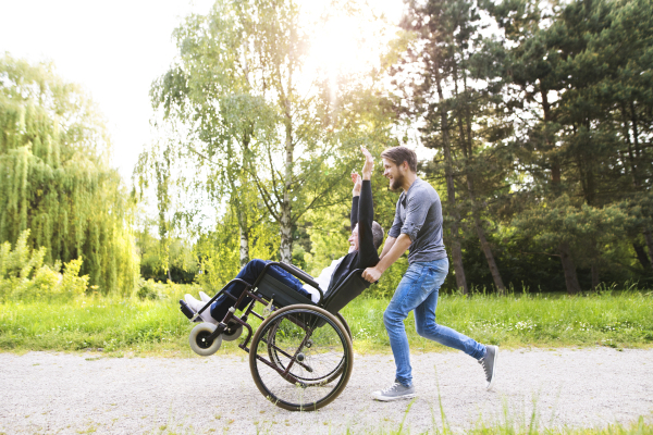 Young hipster son running with disabled father in wheelchair at park, having fun together. Carer assisting disabled senior man.