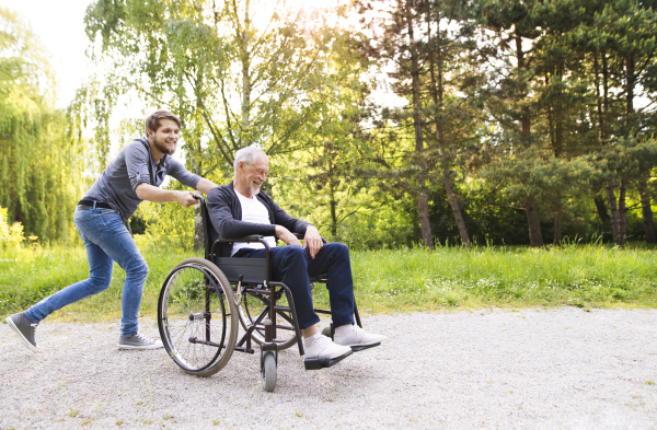 Young hipster son running with disabled father in wheelchair at park, having fun together. Carer assisting disabled senior man.