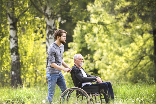 Young hipster son walking with disabled father in wheelchair at park. Carer assisting disabled senior man.