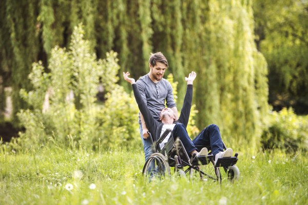 Young hipster son walking with disabled father in wheelchair at park, having fun together. Carer assisting disabled senior man.