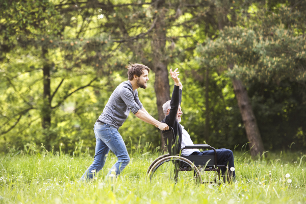 Young hipster son walking with disabled father in wheelchair at park, having fun together. Carer assisting disabled senior man.