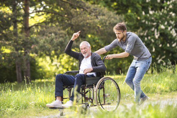 Young hipster son walking with disabled father in wheelchair at park, having fun together. Carer assisting disabled senior man.