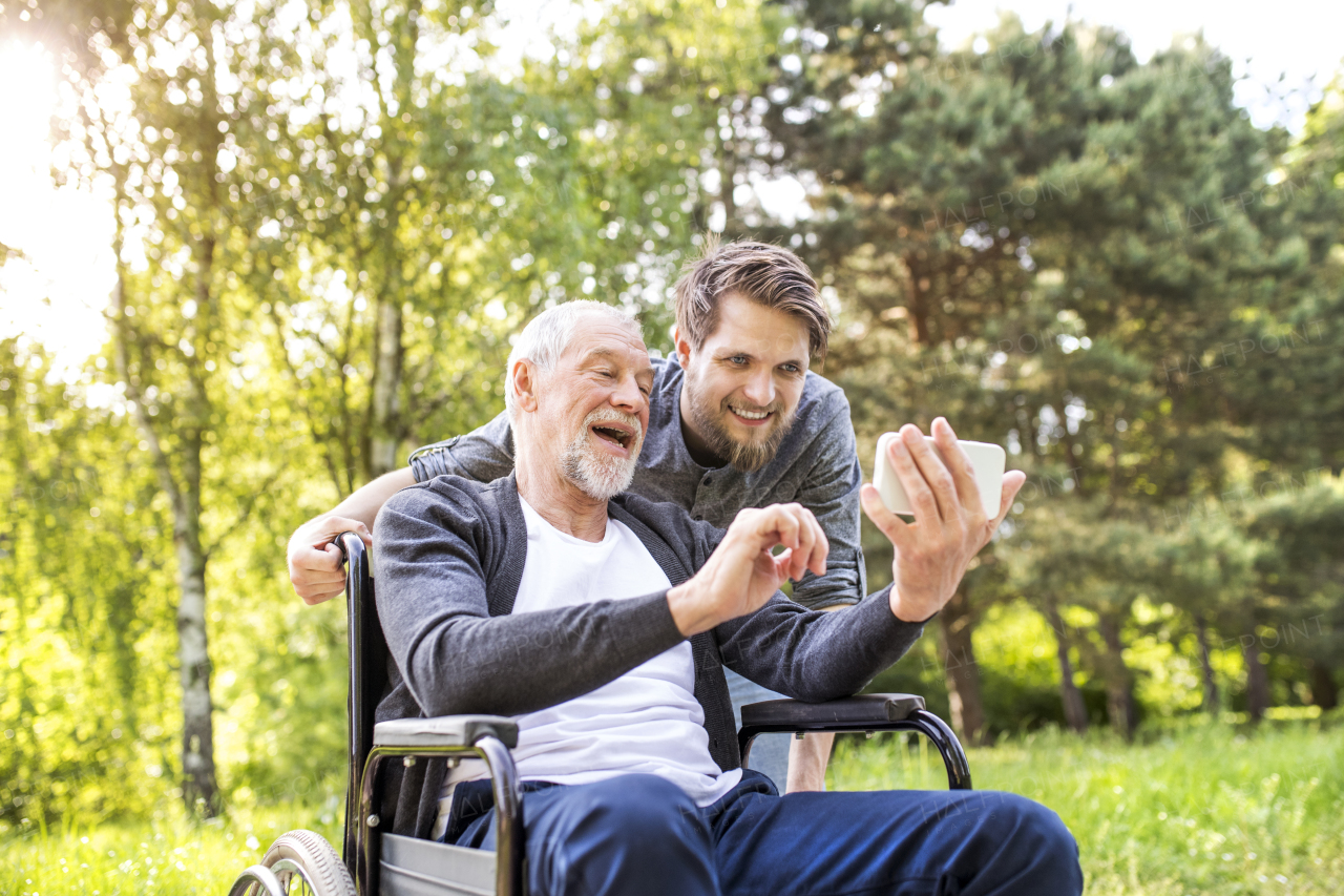 Young hipster son with disabled father in wheelchair at park taking selfie with smart phone. Carer assisting disabled senior man.