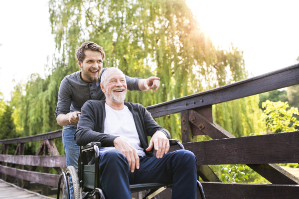 Young hipster son walking with disabled father in wheelchair on wooden bridge at park, pointing finger at something. Carer assisting disabled senior man.