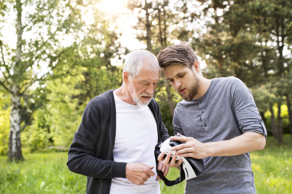 Young man with his senior father in park showing him how does virtual reality glasses work. Sunny summer nature.