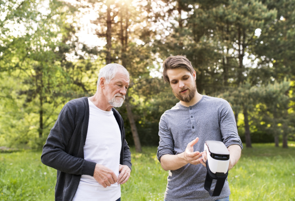 Young man with his senior father in park showing him how does virtual reality glasses work. Sunny summer nature.