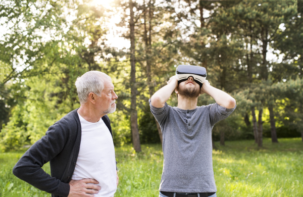 Young man with his senior father in park showing him how does virtual reality glasses work. Sunny summer nature.