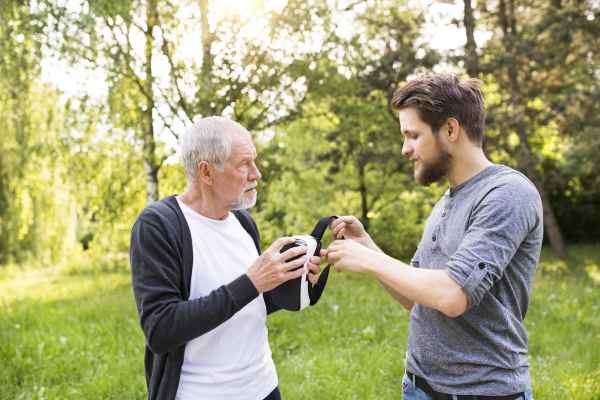 Young man with his senior father in park showing him how does virtual reality glasses work. Sunny summer nature.