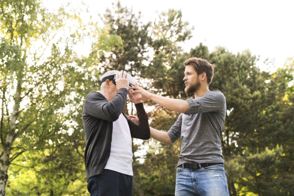 Young man with his senior father in park showing him how does virtual reality glasses work. Sunny summer nature.