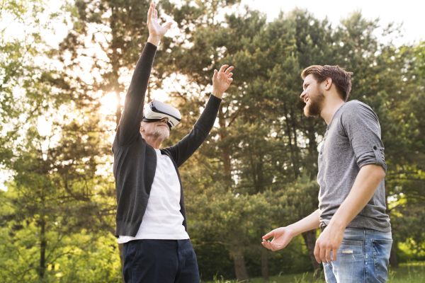 Young man with his senior father in park showing him how to use virtual reality glasses. Sunny summer nature.