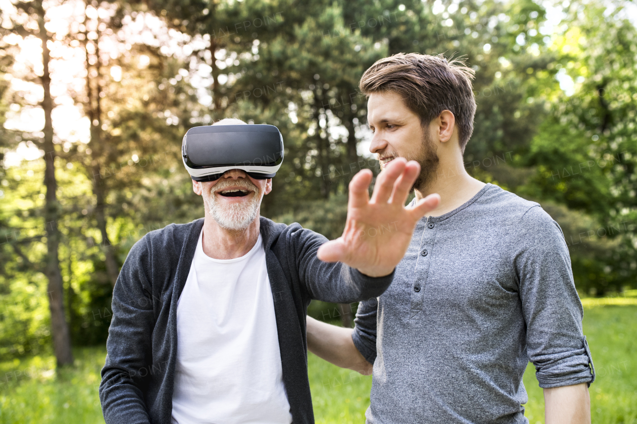 Young man with his senior father in park showing him how to use virtual reality glasses. Sunny summer nature.