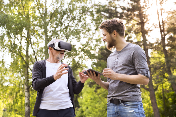 Young man with tablet with his senior father in park showing him how to use virtual reality glasses. Sunny summer nature.