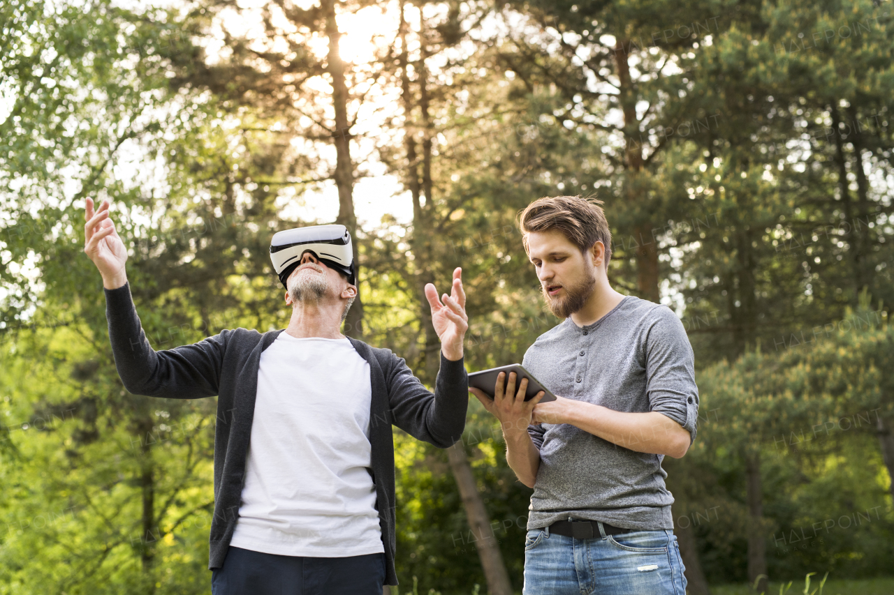 Young man with tablet with his senior father in park showing him how to use virtual reality glasses. Sunny summer nature.