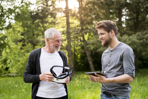 Young man with tablet with his senior father in park showing him how to use virtual reality glasses. Sunny summer nature.