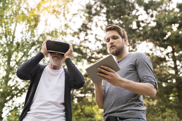 Young man with tablet with his senior father in park showing him how to use virtual reality glasses. Sunny summer nature.