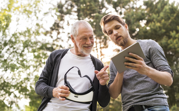 Young man with tablet with his senior father in park showing him how to use virtual reality glasses. Sunny summer nature.