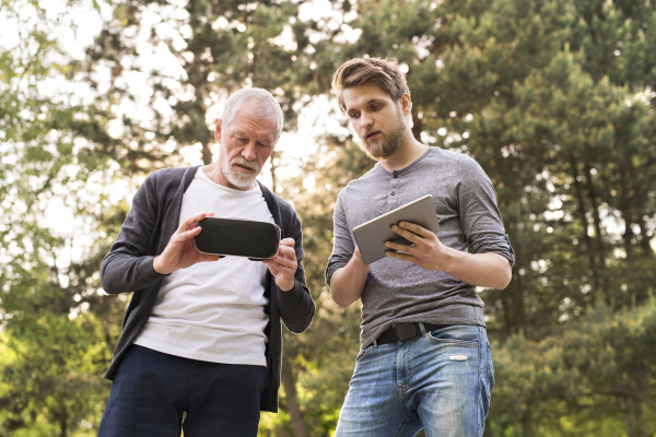 Young man with tablet with his senior father in park showing him how to use virtual reality glasses. Sunny summer nature.