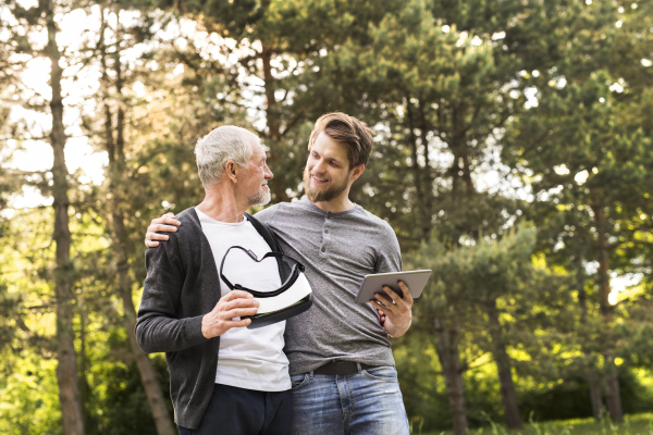 Young man with tablet with his senior father in park showing him how to use virtual reality glasses. Sunny summer nature.