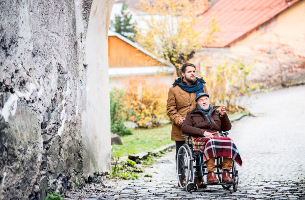 Father in wheelchair and young son on a walk. A carer assisting disabled senior man.