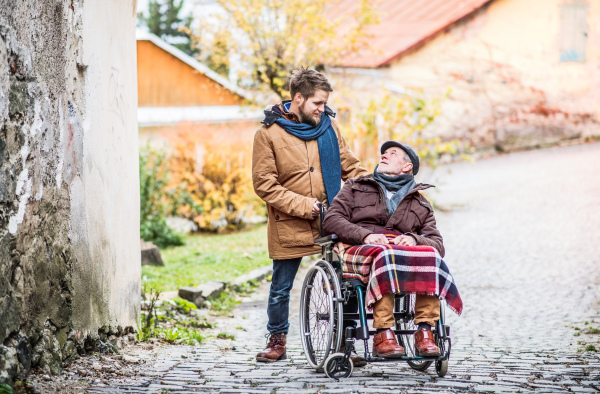 Father in wheelchair and young son on a walk. A carer assisting disabled senior man.