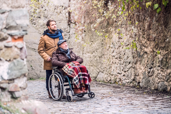 Father in wheelchair and young son on a walk in old town. A carer assisting disabled senior man.