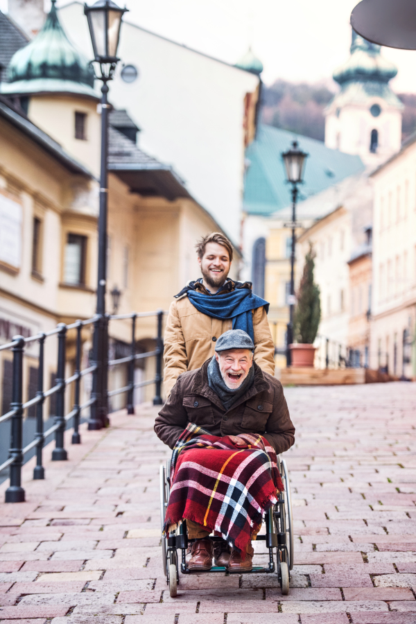 Father in wheelchair and young son on a walk, laughing. A carer assisting disabled senior man.