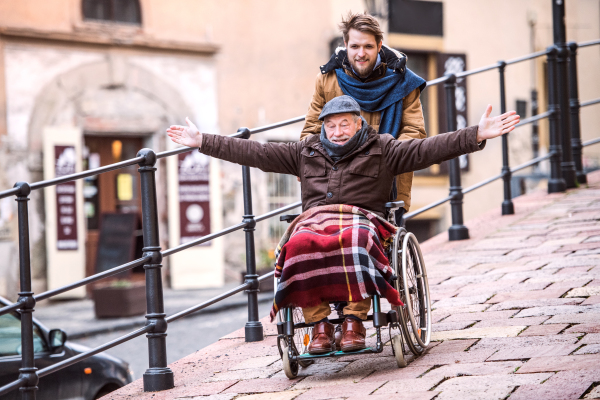 Father in wheelchair and young son on a walk, having fun. A carer assisting disabled senior man.