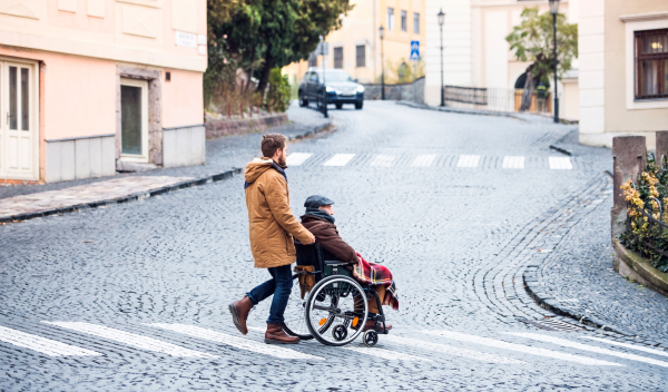 Father in wheelchair and young son on a walk, crossing the road. A carer assisting disabled senior man.