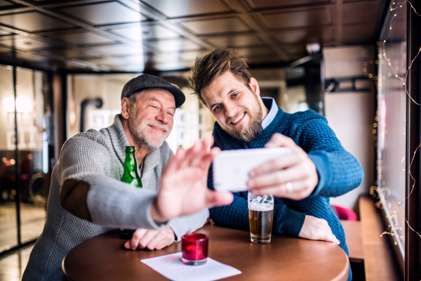 Senior father and his young son drinking beer in a pub. A young man taking selfie with smartphone.